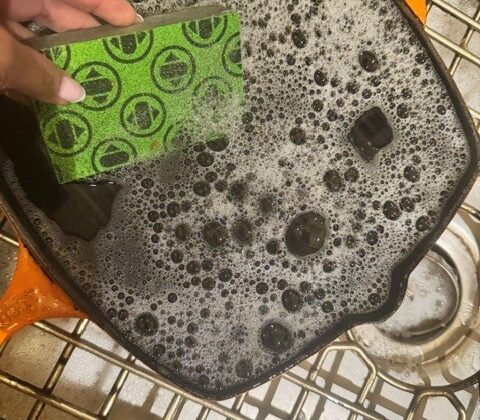 Hand holding a green Skura sponge, its faded dot pattern still visible, scrubbing a sudsy cast iron pan in a sink