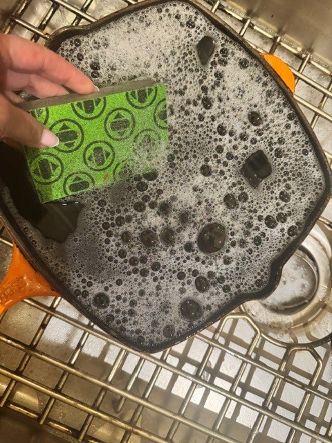 Hand holding a green Skura sponge, its faded dot pattern still visible, scrubbing a sudsy cast iron pan in a sink