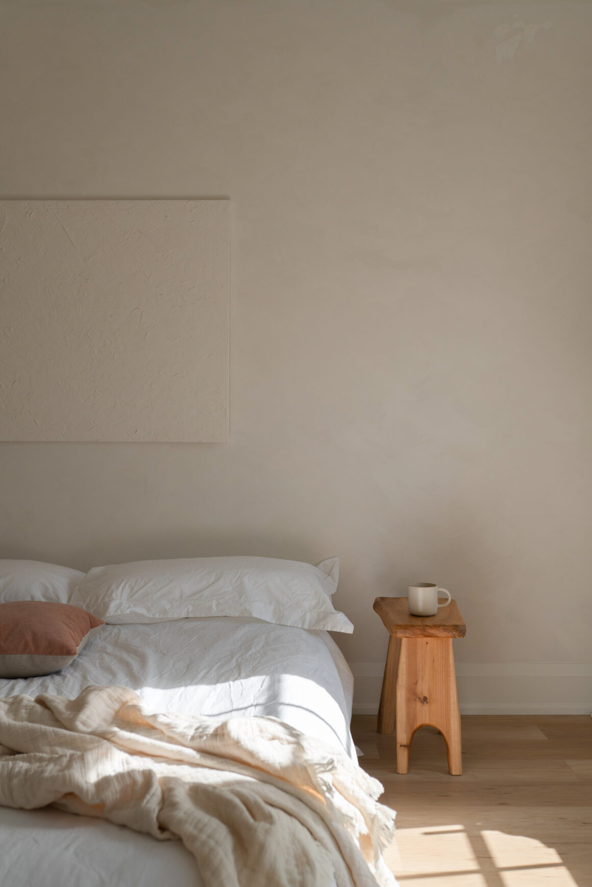 A neatly made bed with white linen bedding and a terracotta pillow, beside a small wooden stool holding a coffee mug, in soft morning light.