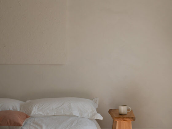A neatly made bed with white linen bedding and a terracotta pillow, beside a small wooden stool holding a coffee mug, in soft morning light.