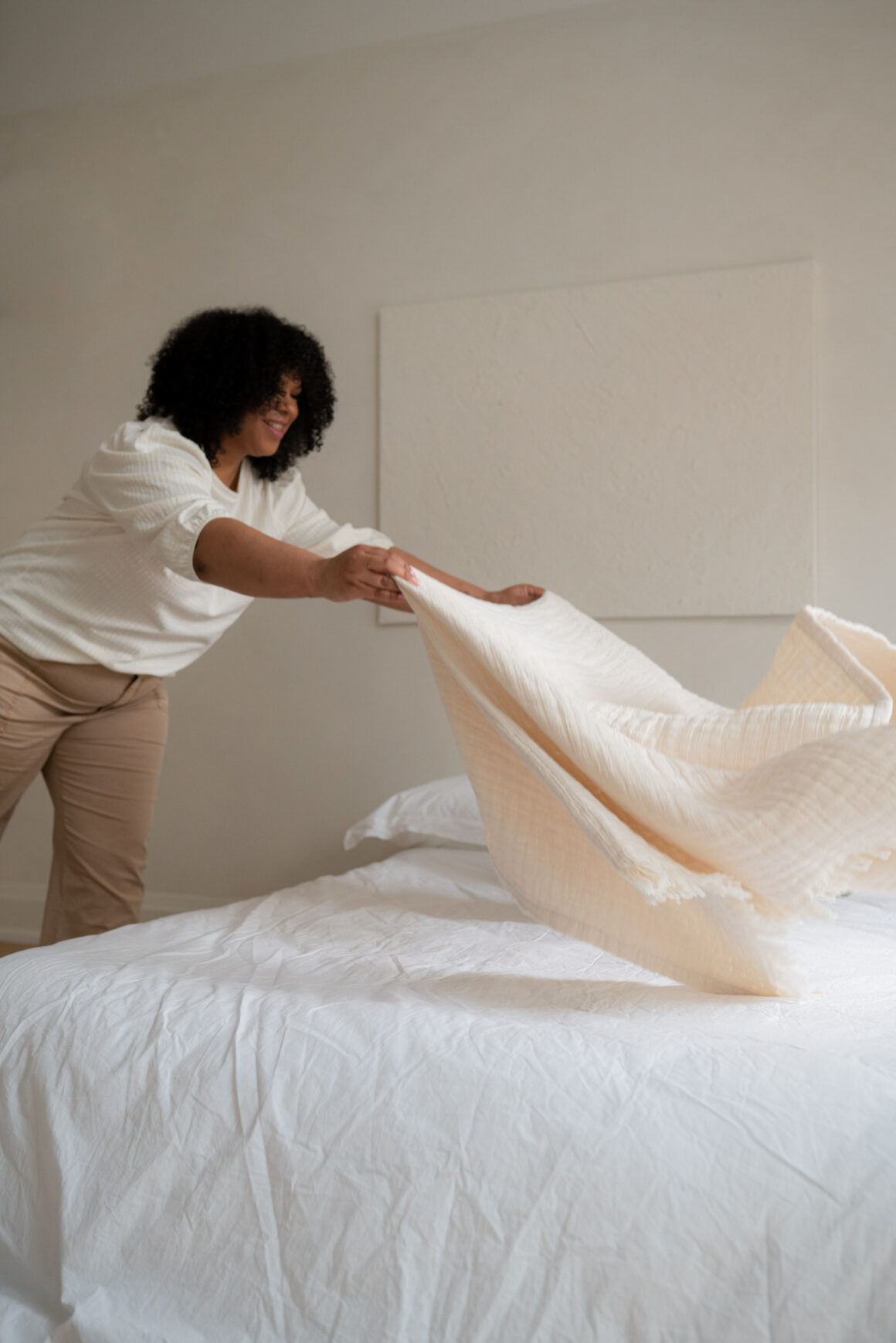 A woman smiling as she spreads a soft throw over fresh white sheets in a bright bedroom — healthkeeping in action.