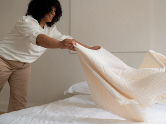A woman smiling as she spreads a soft throw over fresh white sheets in a bright bedroom — healthkeeping in action.
