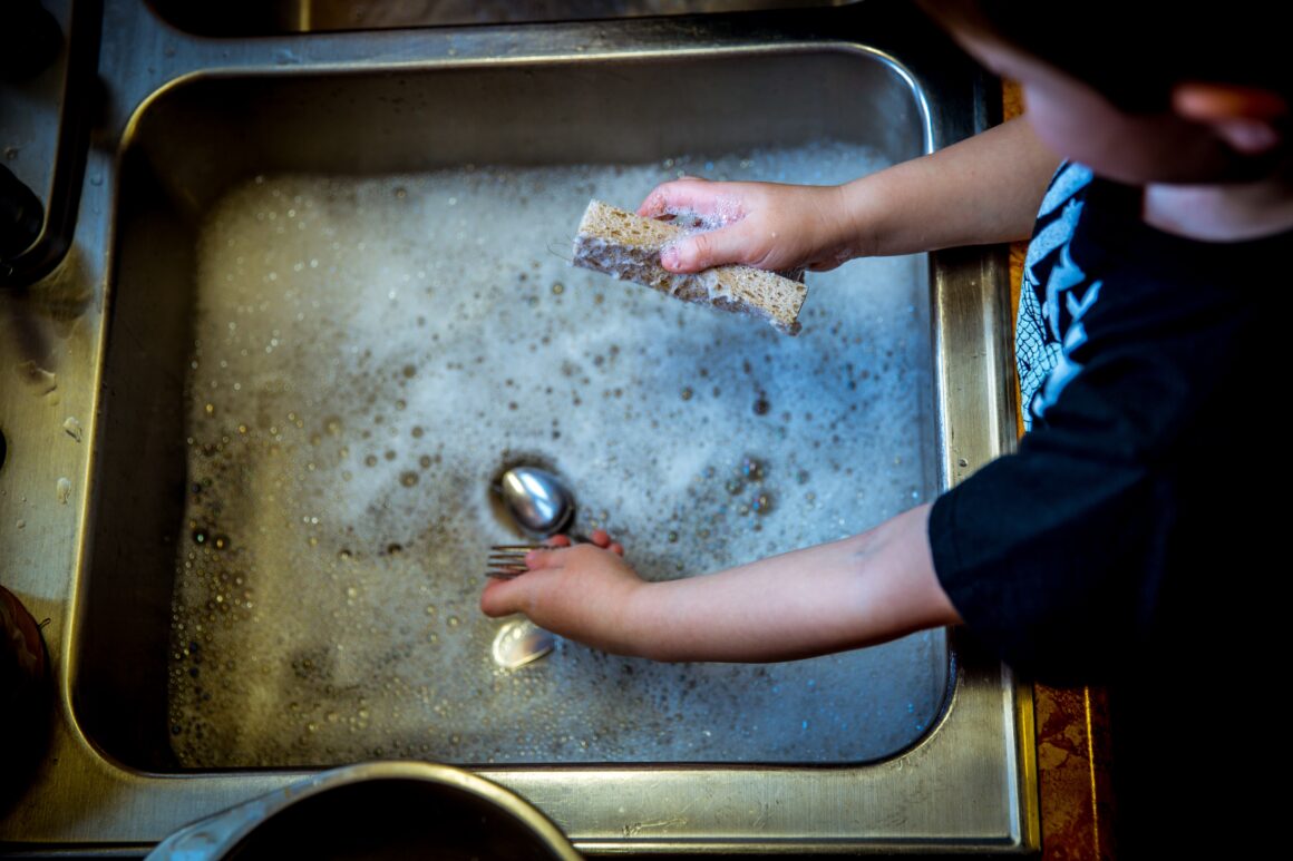 Hands washing a fork and spoon with a worn sponge in a soapy stainless steel sink.