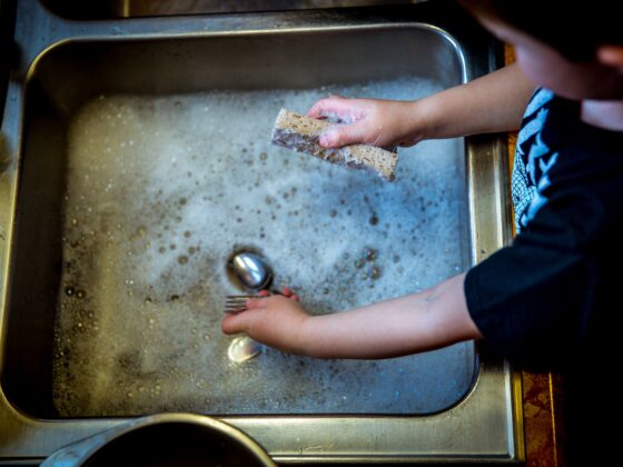 Hands washing a fork and spoon with a worn sponge in a soapy stainless steel sink.