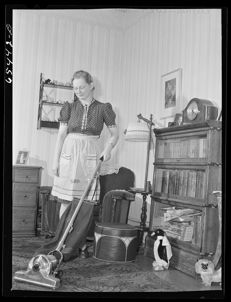 Woman with a vintage upright vacuum cleaner in a wood-paneled living room, the kind that can send dust back into the air.