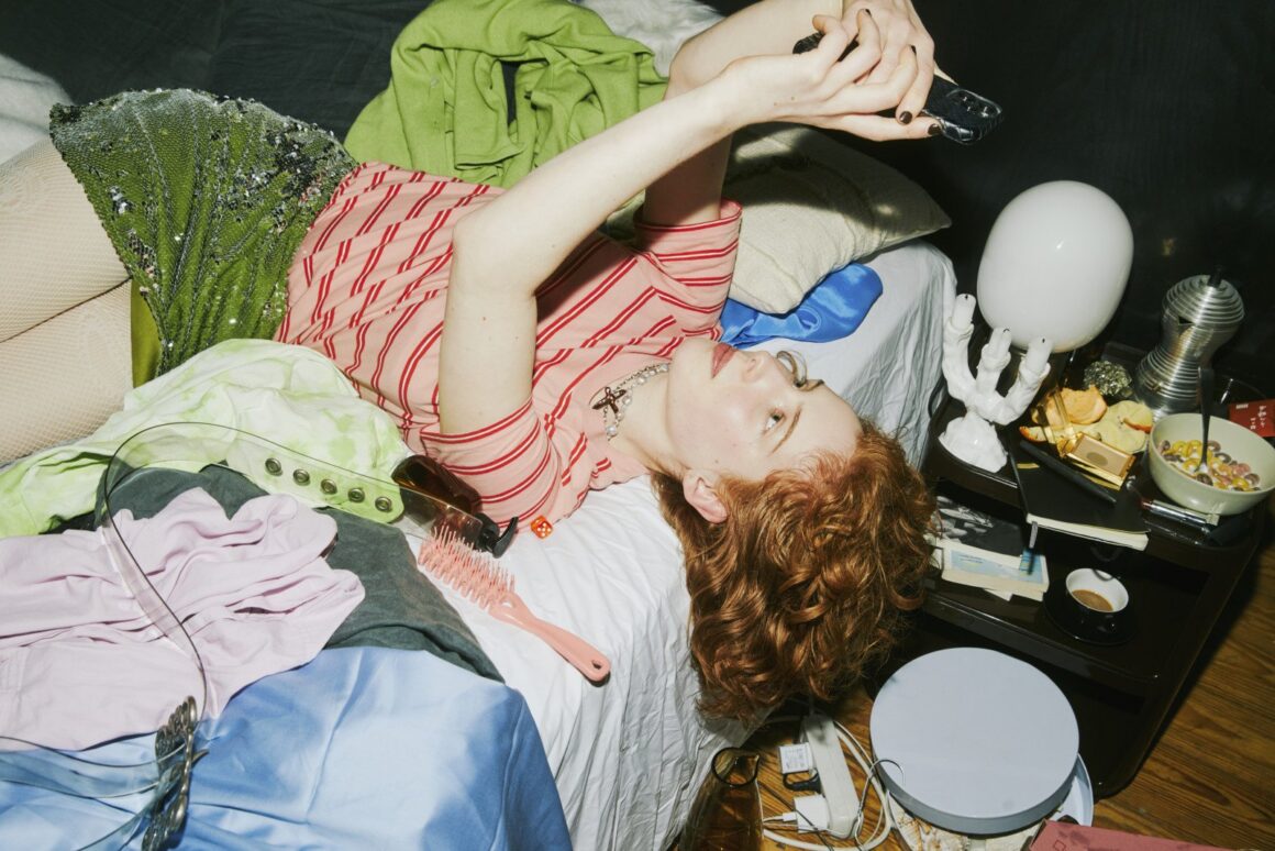 A young woman lies back on her phone, relaxed, surrounded by clothes and clutter in a messy bedroom.