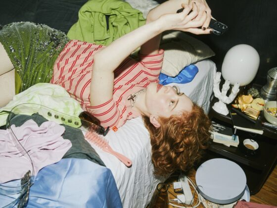 A young woman lies back on her phone, relaxed, surrounded by clothes and clutter in a messy bedroom.