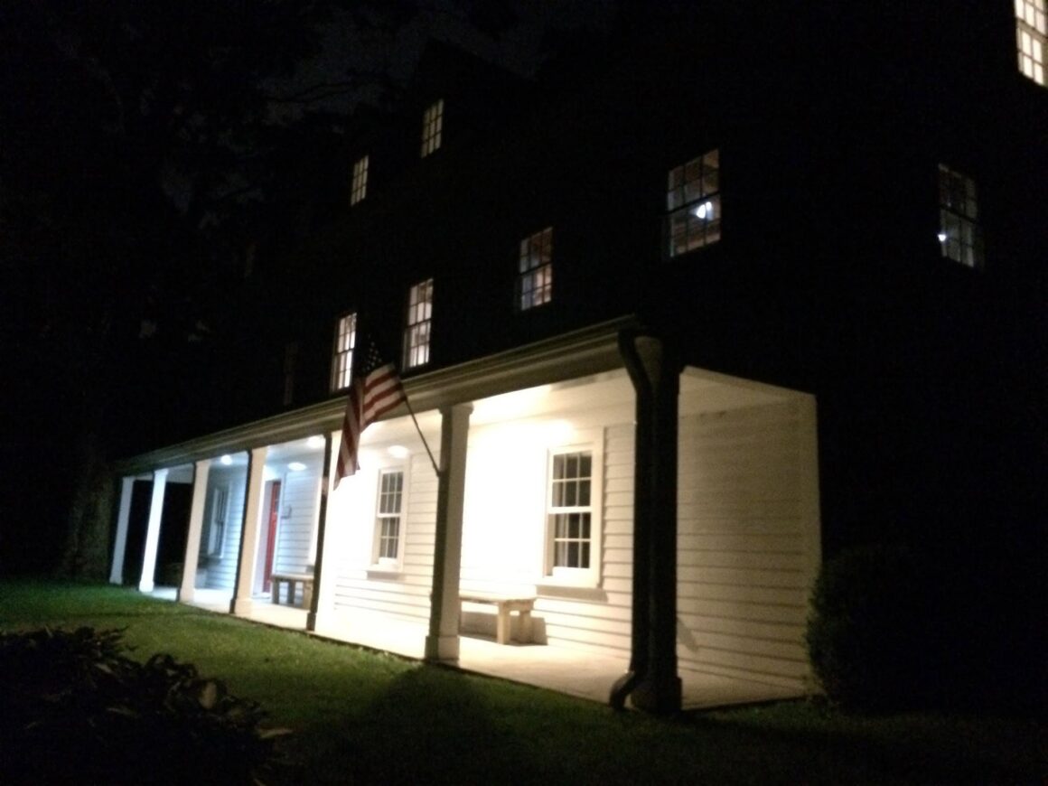 A white colonial house photographed at night, porch lights and several upstairs windows glowing warm, an American flag hanging from a porch column, and a bench on the covered porch.