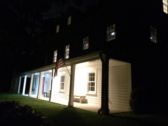 A white colonial house photographed at night, porch lights and several upstairs windows glowing warm, an American flag hanging from a porch column, and a bench on the covered porch.