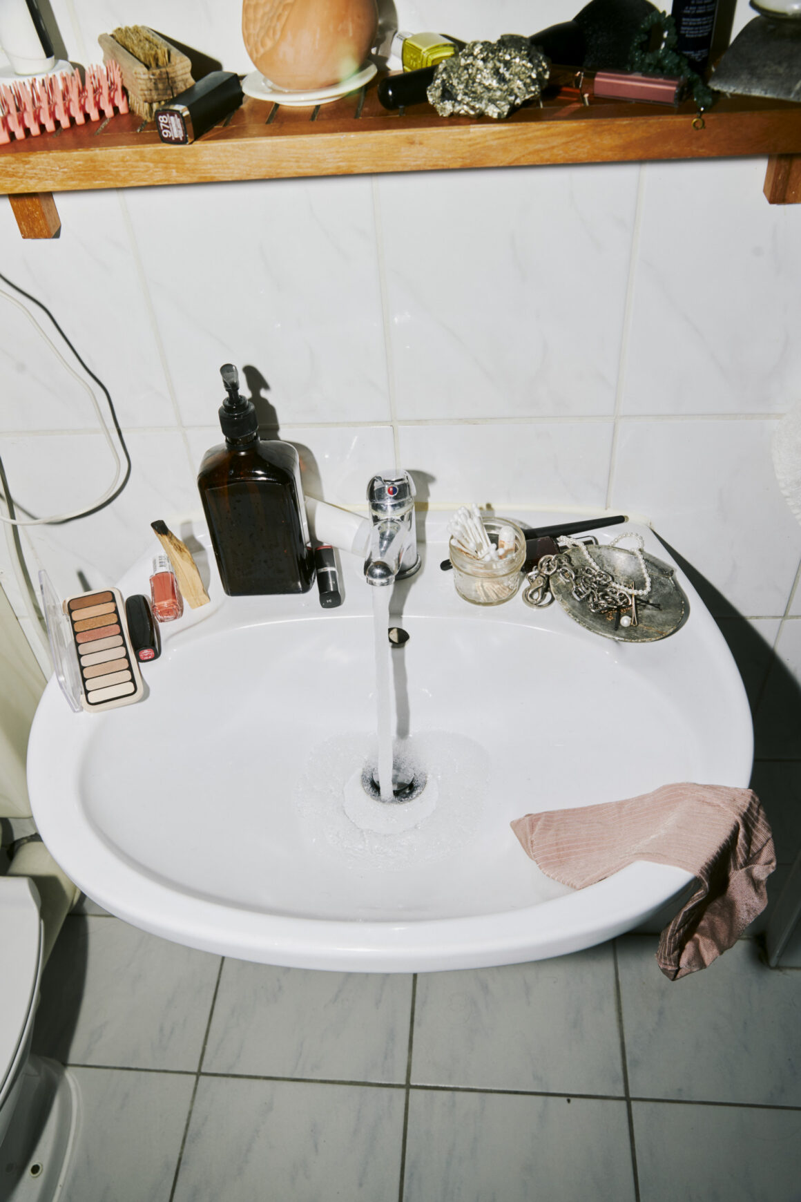 A bathroom sink with the tap running, a towel draped over one edge, and a shelf of everyday toiletries above it.