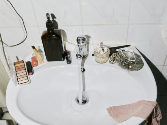 A bathroom sink with the tap running, a towel draped over one edge, and a shelf of everyday toiletries above it.