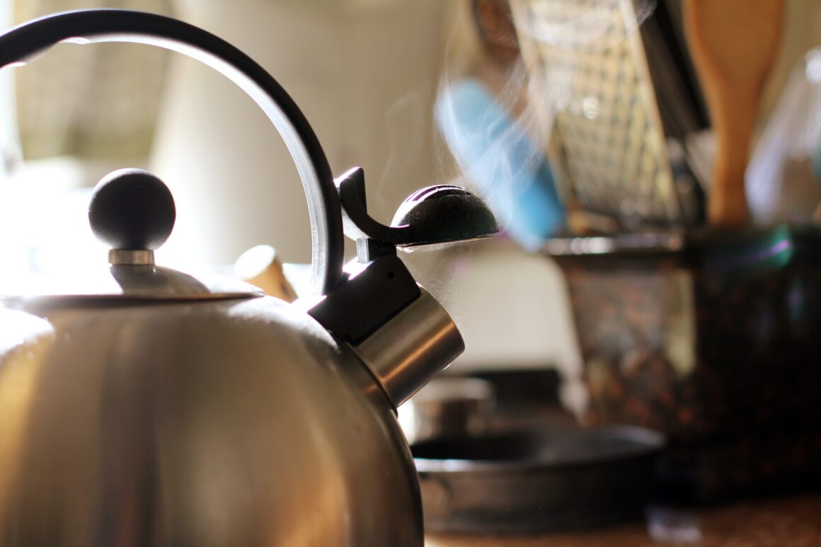 A stainless steel kettle steaming on a stovetop in a sunlit kitchen