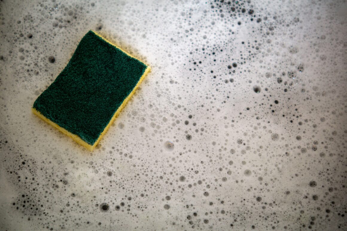 Green and yellow scrub sponge resting in soapy water on a gray surface.