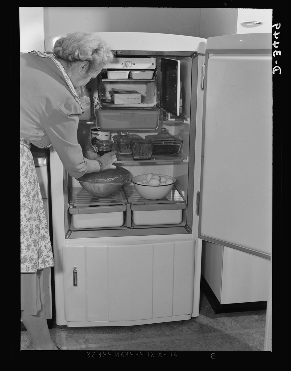 A white-haired woman in an apron leans into an open vintage refrigerator stocked with glass dishes, jars, a bottle of milk, a box of frozen peas, and a bowl of eggs.