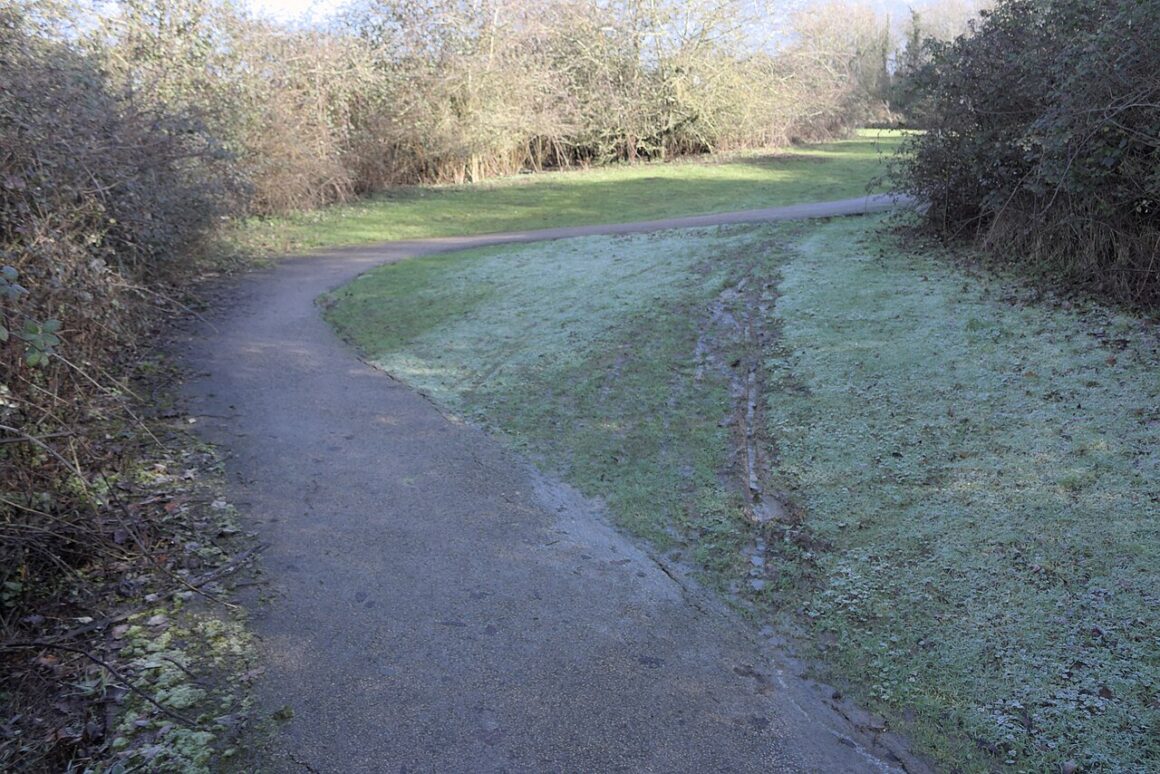 A muddy shortcut worn across grass beside a curving paved [atj