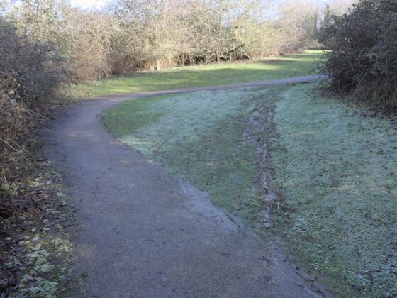 A muddy shortcut worn across grass beside a curving paved [atj