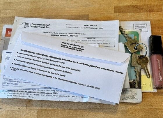 A stack of mail on a wooden counter, topped by a New York State DMV license renewal notice, with car keys and a tube of lip gloss beside it.