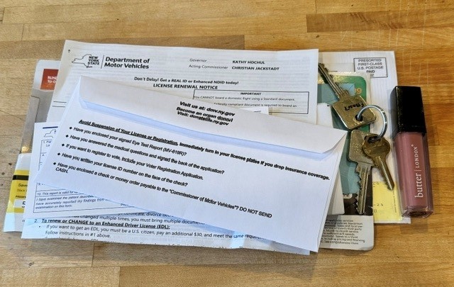 A stack of mail on a wooden counter, topped by a New York State DMV license renewal notice, with car keys and a tube of lip gloss beside it.