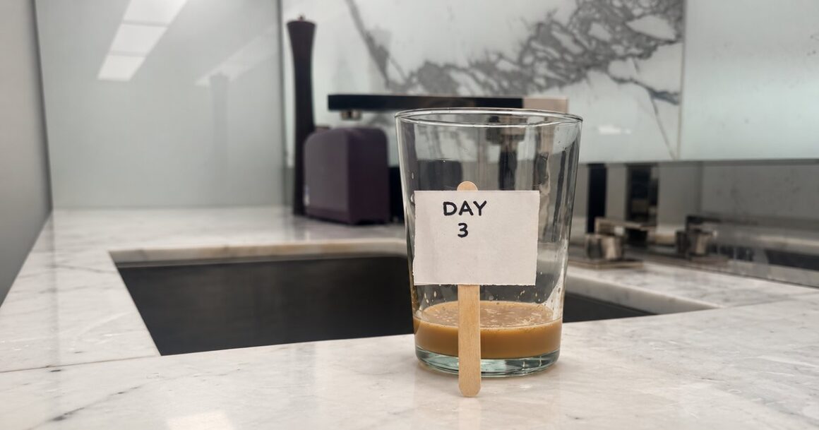 A glass with an inch of leftover coffee on a kitchen counter next to the sink, with a small handwritten sign on a popsicle stick that reads "Day 3.