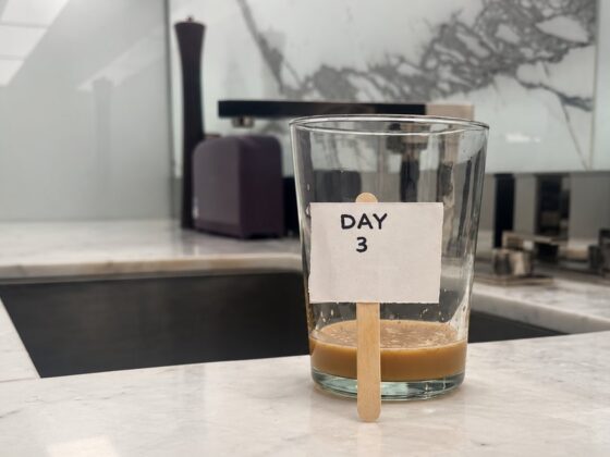 A glass with an inch of leftover coffee on a kitchen counter next to the sink, with a small handwritten sign on a popsicle stick that reads "Day 3.
