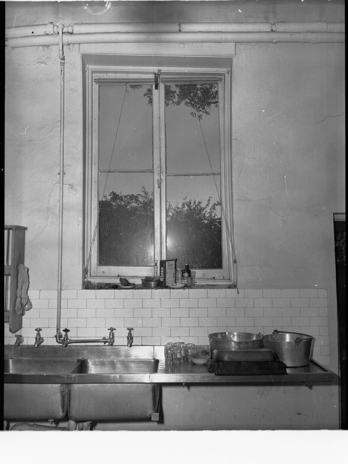 Black and white photo of a kitchen sink beneath a casement window at Government House, Adelaide, with subway tile and exposed pipes