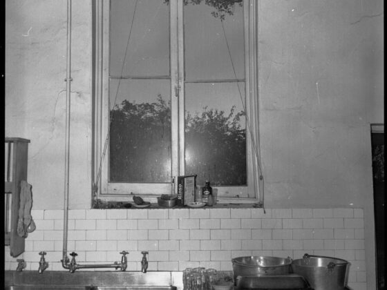 Black and white photo of a kitchen sink beneath a casement window at Government House, Adelaide, with subway tile and exposed pipes