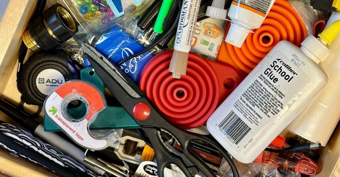 A cluttered wooden kitchen drawer with scissors, an orange collapsible funnel, school glue, tape and assorted small tools