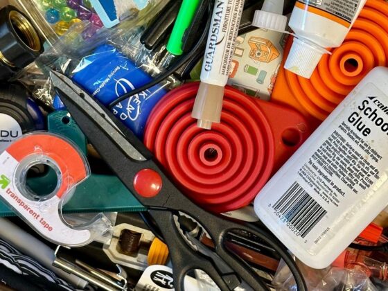 A cluttered wooden kitchen drawer with scissors, an orange collapsible funnel, school glue, tape and assorted small tools