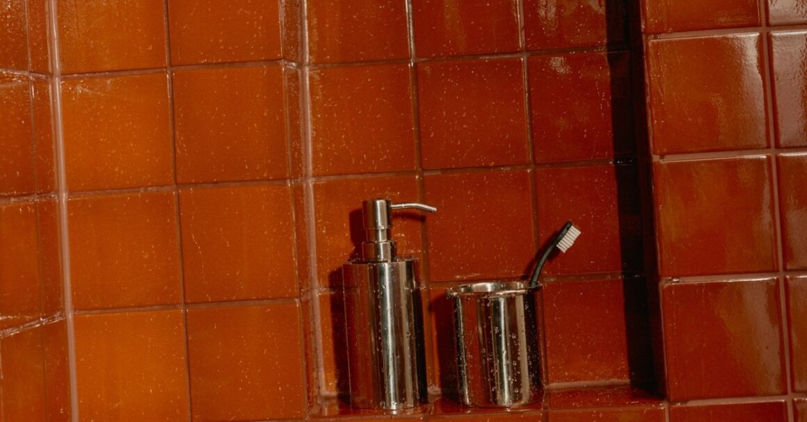 Glossy terracotta shower tile with water droplets and a chrome soap dispenser in a tiled niche